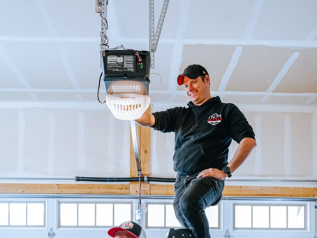 An installer up a step ladder with his hands on the motor head above a garage door, a second man watching from the floor.