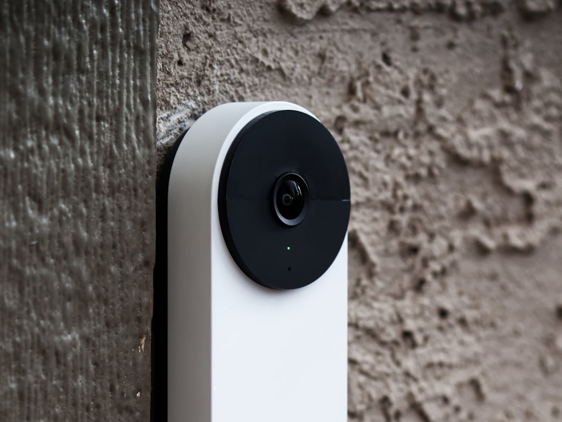A video door station mounted on a plastered wall, its camera lens facing the approach.