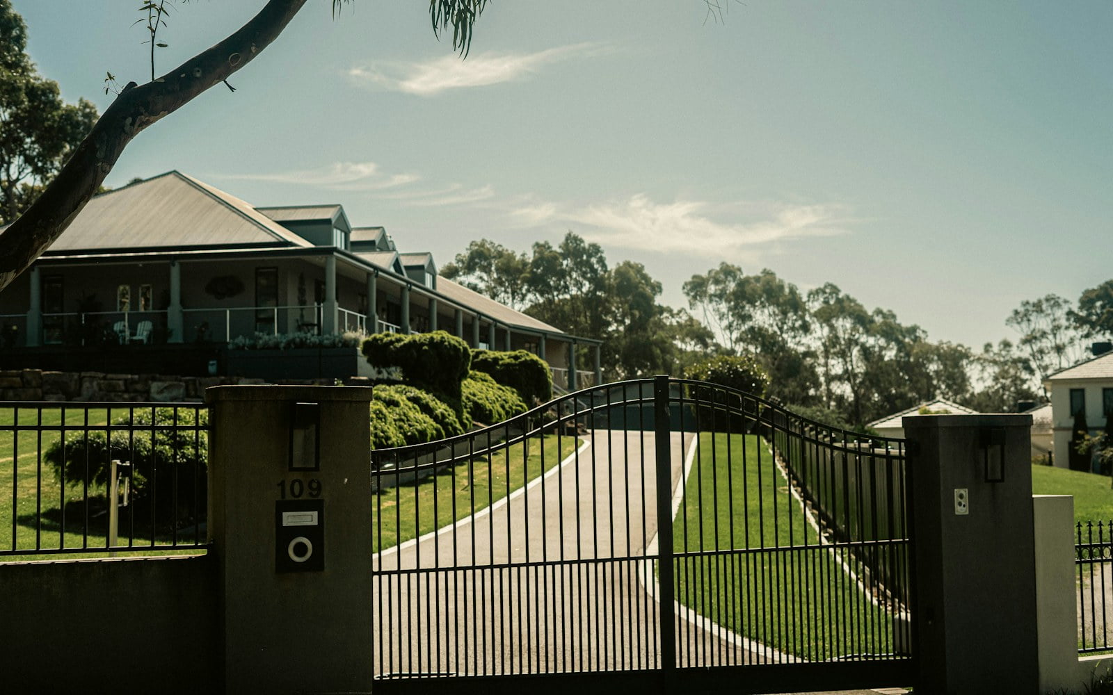 An automated driveway gate standing open, with the intercom post on the kerb beside it and the house up the drive behind.