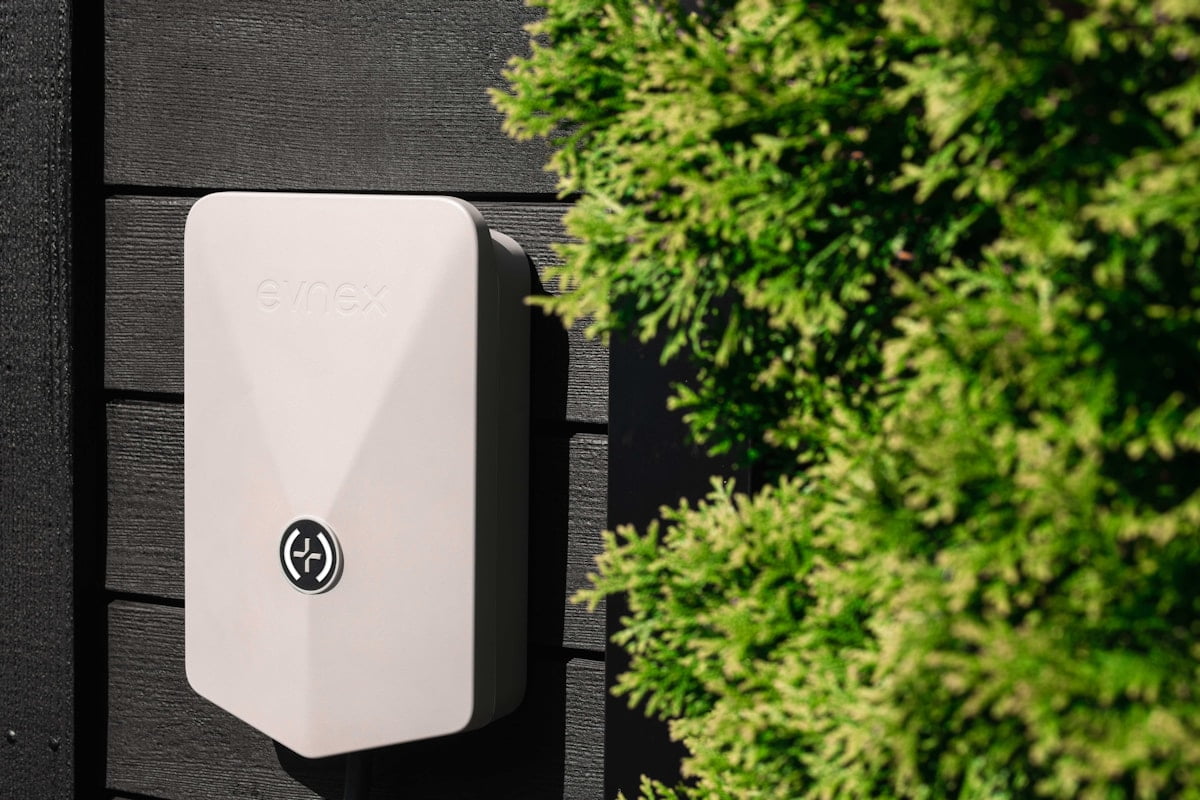 A weatherproof intercom station fixed to a dark exterior wall beside a hedge.